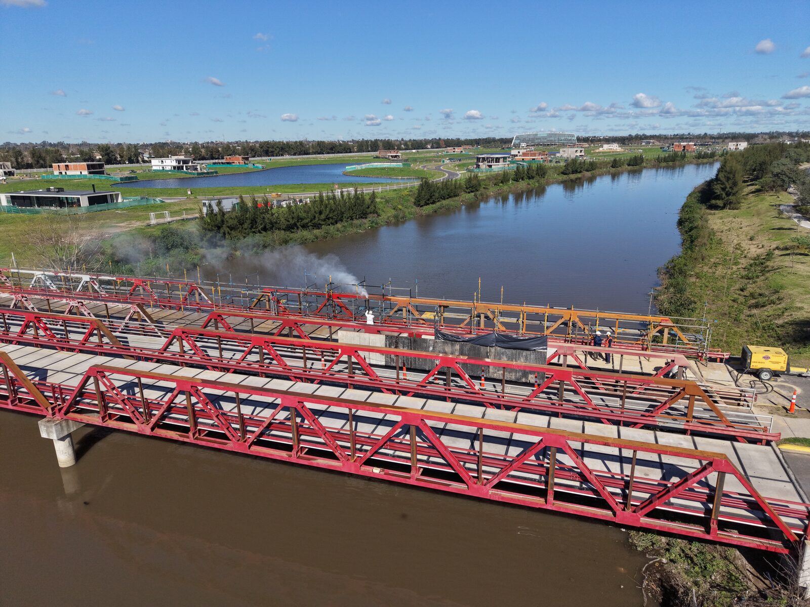 Vista aérea del puente metálico rojo sobre el arroyo, con el equipo trabajando en el arenado de la estructura.